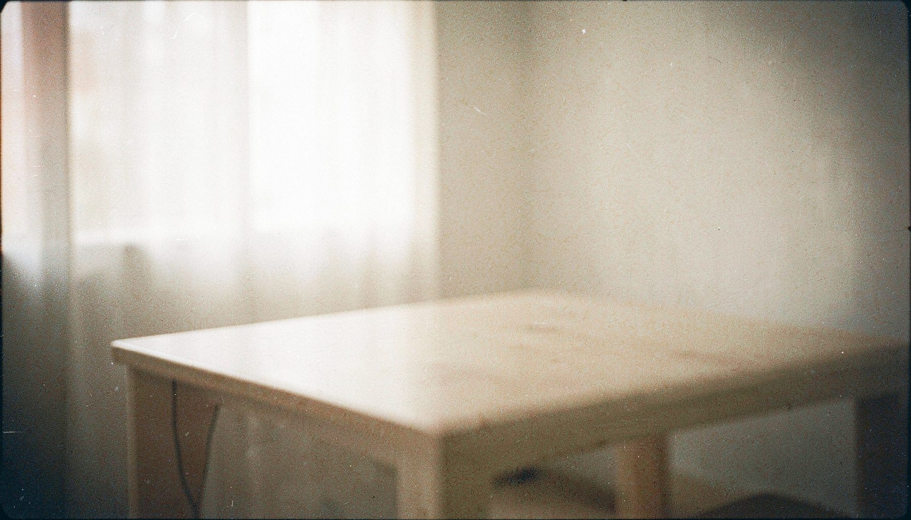 A grainy analog film photograph of a minimalist interior with soft beige and off-white tones, an empty tabletop, and diffused window light creating a serene, neutral mood.