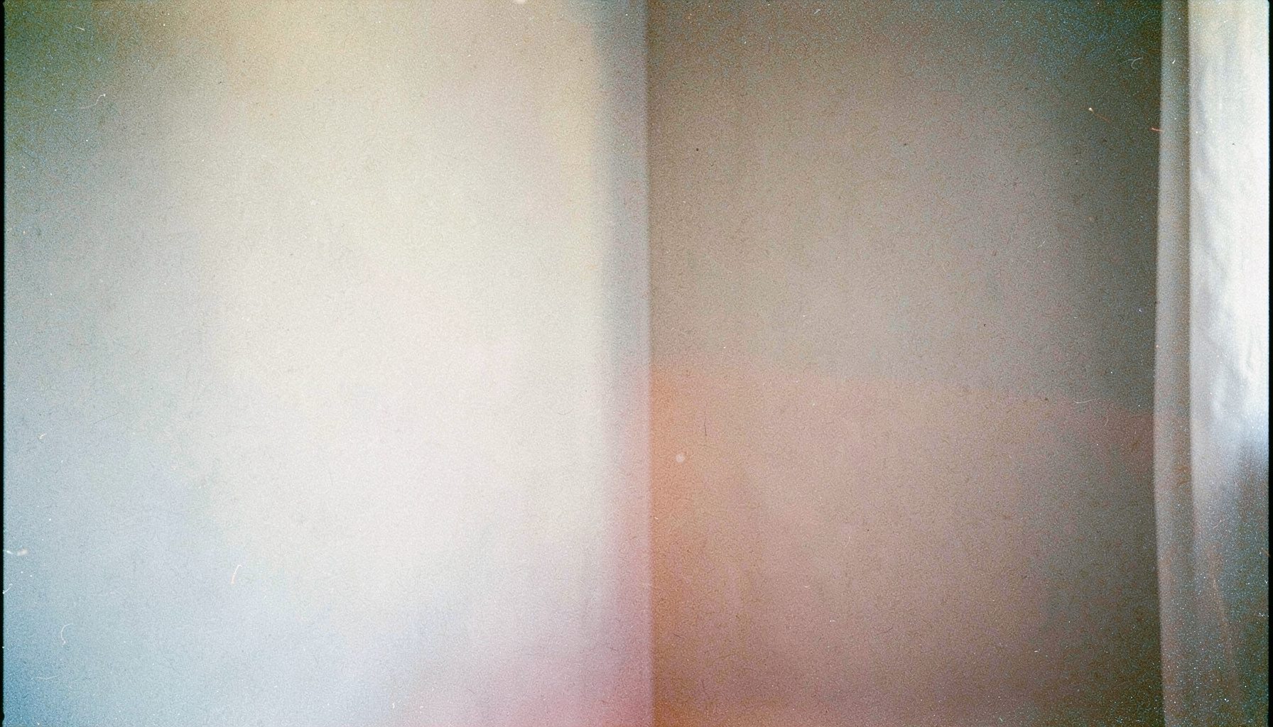 A grainy analog film photograph of a pale pastel-toned room corner with a soft pink accent, muted blue-grey shadows, and warm natural light, conveying a calm and welcoming feel.