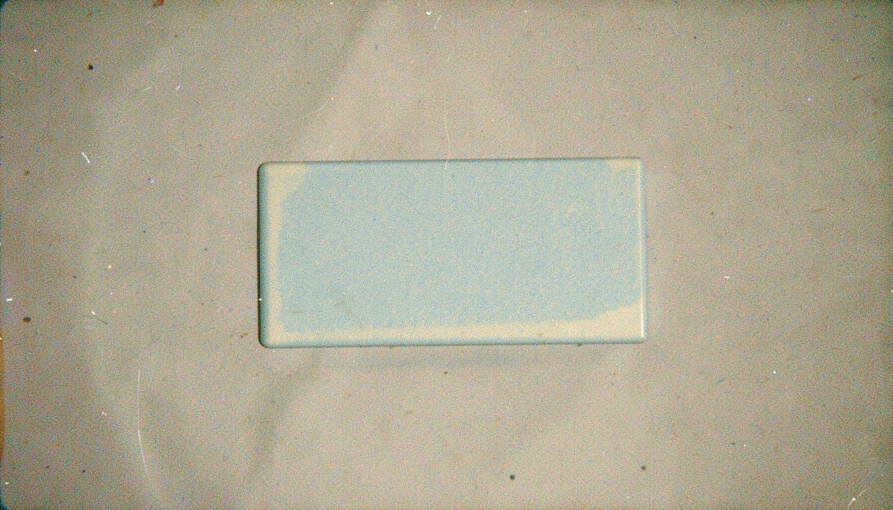 A grainy analog film photograph of a small rectangular badge with muted pastel blue and cream tones, subtle and minimal.