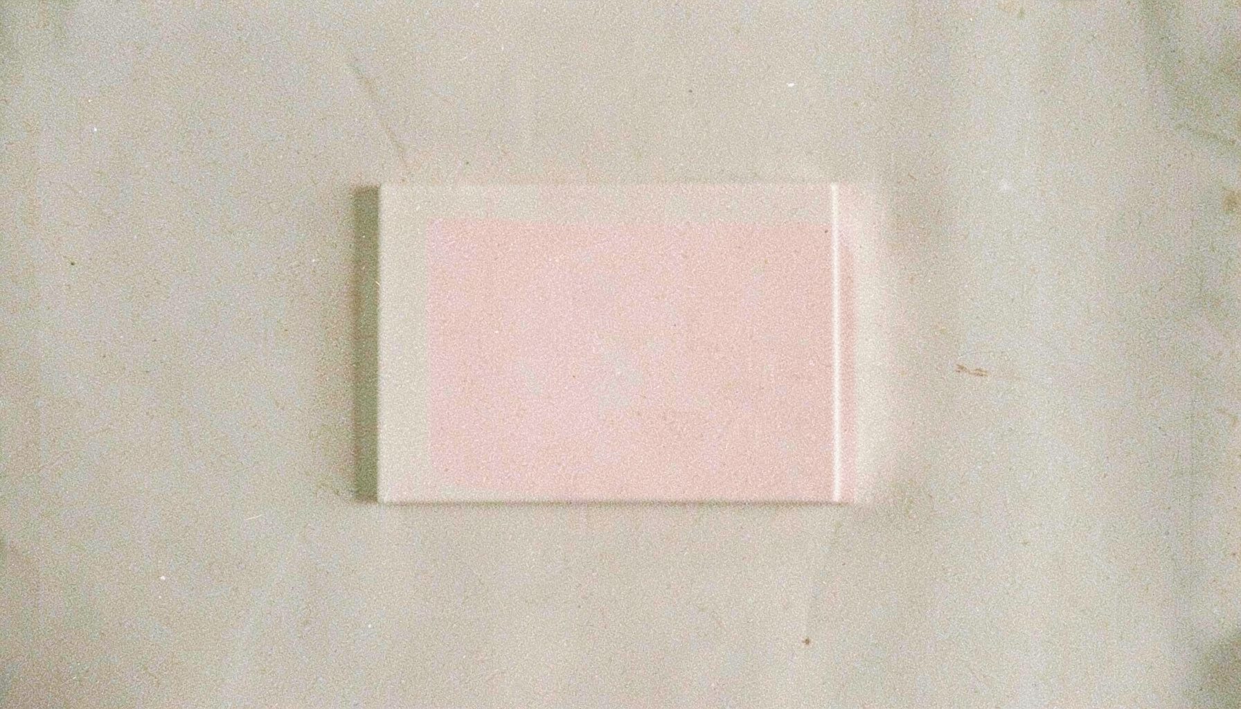 A grainy analog film photograph of a small rectangular badge with soft pastel pink and off-white tones, understated and clean.