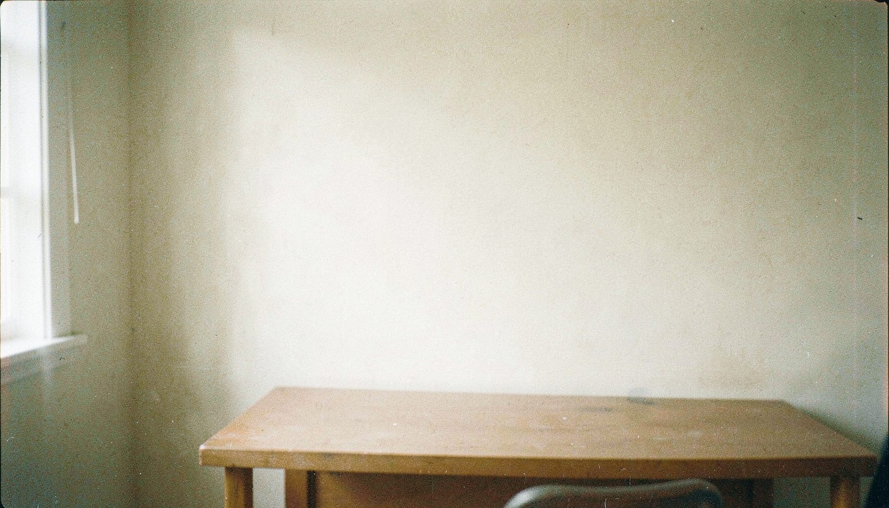 A soft, grainy analog film photograph of a calm, neutral-toned workspace with a pale cream wall, a simple wooden desk surface, and gentle natural light casting faint shadows, evoking a quiet and understated atmosphere.